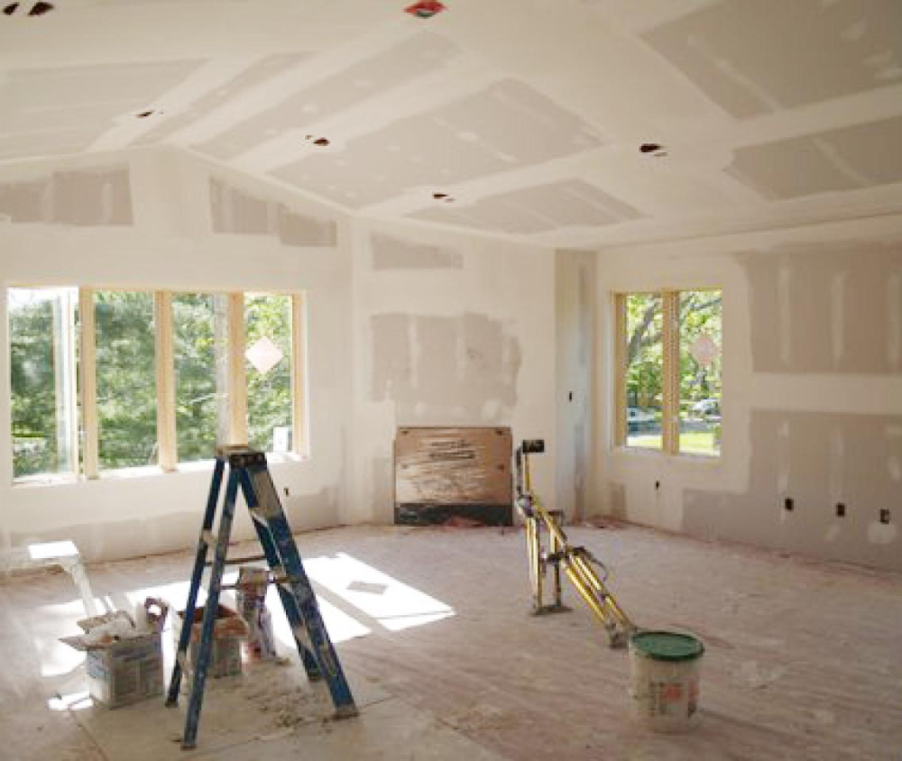 A newly drywalled room with a vaulted ceiling, recessed lighting, a bank of windows, and a rough opening, mid finish before paint.