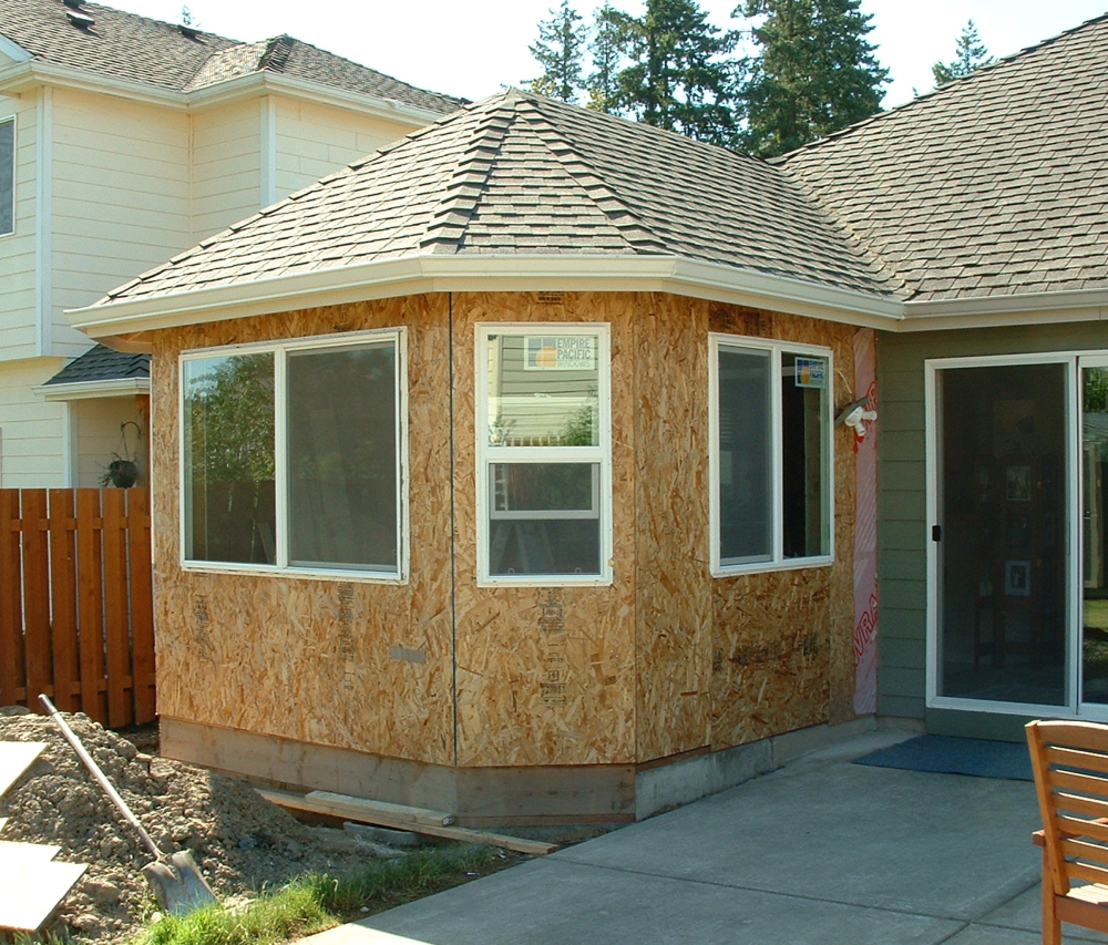 A hip-roofed addition sheathed in OSB with white-framed window openings and a sliding glass door, tied into the existing house on a concrete foundation.